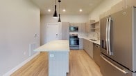 This single-floor residence showcases a contemporary design characterized by clean lines, a neutral color palette, and light hardwood-style flooring throughout. The open-concept kitchen serves as the centerpiece of the living area, featuring flat-front cabinetry in a warm light wood finish, white quartz countertops, and a full suite of stainless steel appliances including a French door refrigerator, dishwasher, and range with microwave. A spacious central island with a matching quartz surface provides additional workspace and casual seating, complemented by striking pendant lighting overhead. Recessed lighting and additional pendant fixtures ensure the kitchen and living area are well-illuminated. The layout, as seen from the overhead view, flows seamlessly from the kitchen toward the living space, which opens to a balcony through sliding glass doors, enhancing the sense of openness. The bedroom offers a tranquil retreat, with a large window framing views of autumn foliage and the surrounding cityscape, flooding the room with natural light. The bathroom is finished with the same cohesive aesthetic, featuring a light wood vanity with quartz countertop, a large mirror, and a full tub-shower combination. Overall, the space presents a bright, modern, and functional living environment.