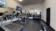 Located at 801 Olympic Square in Aberdeen, Maryland, this single-floor fitness center offers a comprehensive workout environment designed to support a wide range of exercise routines. The space features durable rubber flooring throughout, providing both safety and comfort during workouts.

The cardio zone is equipped with multiple treadmills and an elliptical machine, each positioned for easy use and accompanied by wall-mounted flat-screen televisions to keep members entertained during their sessions. Full-length mirrors line one wall, visually expanding the space and allowing users to monitor their form.

The strength training area includes a well-stocked dumbbell rack with a variety of weight options, several cable and resistance machines for targeted muscle work, and dedicated leg and arm machines positioned along a windowed wall that brings in natural light from the exterior. Additional functional training equipment such as a cable pull system with attachments rounds out the strength section.

Motivational wall decals reading "The Body Achieves What The Mind Believes" and "Everyday Is A Good Day To Work Out" add an uplifting atmosphere to the space. A small storage bench and sanitation station are also available near the entrance for member convenience. Bright overhead fluorescent lighting ensures the entire facility is well-lit at all times.