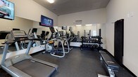Located at 801 Olympic Square in Aberdeen, Maryland, this single-floor fitness center offers a comprehensive workout environment designed to support a wide range of exercise routines. The space features durable rubber flooring throughout, providing both safety and comfort during workouts.

The cardio zone is equipped with multiple treadmills and an elliptical machine, each positioned for easy use and accompanied by wall-mounted flat-screen televisions to keep members entertained during their sessions. Full-length mirrors line one wall, visually expanding the space and allowing users to monitor their form.

The strength training area includes a well-stocked dumbbell rack with a variety of weight options, several cable and resistance machines for targeted muscle work, and dedicated leg and arm machines positioned along a windowed wall that brings in natural light from the exterior. Additional functional training equipment such as a cable pull system with attachments rounds out the strength section.

Motivational wall decals reading "The Body Achieves What The Mind Believes" and "Everyday Is A Good Day To Work Out" add an uplifting atmosphere to the space. A small storage bench and sanitation station are also available near the entrance for member convenience. Bright overhead fluorescent lighting ensures the entire facility is well-lit at all times.