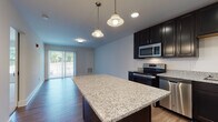 This single-floor unit in Groton, Connecticut offers a thoughtfully designed layout with modern finishes throughout. The kitchen serves as a centerpiece of the space, showcasing rich dark cabinetry that extends floor-to-ceiling, providing generous storage. Granite countertops with a light speckled finish complement the full suite of stainless steel appliances, including a refrigerator, electric range, built-in microwave, and dishwasher. A substantial kitchen island with matching granite adds both prep space and visual impact, while pendant lighting overhead adds a warm, stylish touch. The kitchen opens naturally toward a living area with wood-look flooring, and sliding glass doors bring in natural light and provide access to an outdoor space with greenery views. The bedroom features plush carpeting, clean white walls, and a wide window that allows for ample natural light. A stylish barn-style sliding door adds a contemporary architectural detail, leading to a well-sized walk-in closet fitted with wire shelving for organized storage. The bathroom is finished with classic black-and-white hexagon tile flooring, a dark vanity with a white countertop, and a full tub-shower combination. Throughout the unit, the design balances modern aesthetics with functional, everyday living details.