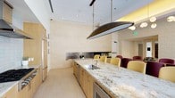 This single-floor space showcases a thoughtfully designed kitchen and dining area that blends contemporary aesthetics with functional elegance. The kitchen is anchored by a generous island finished in white and gold granite, complete with an undermount stainless steel sink and ample seating along one side. Light wood cabinetry runs the full length of the space, complemented by panel-ready refrigeration that maintains a seamless, integrated look. A distinctive oval bronze pendant light hangs above the island, serving as a bold focal point against the bright white ceiling equipped with recessed lighting.

The walls are clad in large-format, cream-toned tile that lends a refined, uniform appearance to the space. A decorative dark subway tile backsplash runs horizontally along the back wall, adding textural contrast. Adjacent to the kitchen, a dining area is furnished with yellow and burgundy upholstered chairs, introducing warm color accents. The light-toned flooring throughout contributes to an open, airy ambiance. Overall, the space presents a polished, modern environment well-suited for both casual and formal gatherings.