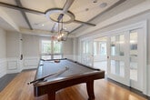 This expansive single-floor amenity space is thoughtfully designed to accommodate a variety of social and recreational activities. The interiors reflect a contemporary aesthetic, blending coffered and beam-accented ceilings with warm hardwood and tile flooring across distinct functional zones.

The main lounge area serves as the centerpiece, featuring a large wall-mounted flat-screen TV set against a textured stone accent wall. Built-in shelving with cabinetry lines one side of the room, complemented by cushioned window bench seating that frames views of the surrounding greenery. Flexible seating arrangements with upholstered ottomans and accent tables create an inviting, versatile gathering space.

Adjacent, a dedicated billiards room offers a full-sized pool table beneath a geometric pendant light fixture, with white-paneled wainscoting and French doors that allow natural light to filter in from multiple directions.

The kitchen area is well-appointed with dark granite countertops, a large central island with bar seating, a mosaic tile backsplash, multiple wall-mounted screens, and lantern-style pendant lighting overhead.

Additional seating nooks feature built-in window benches and casual café-height tables. A tastefully decorated corridor with framed artwork connects the various areas, contributing to the cohesive and polished ambiance throughout.