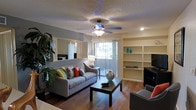This single-floor space offers a thoughtfully designed layout centered around a welcoming living area. The room features rich hardwood-style flooring that adds warmth and visual continuity throughout. A ceiling fan with integrated lighting fixture anchors the space from above, complemented by recessed lighting and a textured ceiling that adds subtle depth.

The living area is generously proportioned, accommodating a plush sofa adorned with colorful accent pillows and a substantial dark wood coffee table. Built-in shelving units line one wall, providing both functional storage and display space, adding an architectural element that enhances the room's character. A large sliding glass door floods the space with natural light and provides direct access to an outdoor balcony, creating a seamless indoor-outdoor connection.

Adjacent to the living area, a mirrored wall with louvered closet doors is visible, reflecting light and amplifying the sense of space. Lush indoor greenery adds a natural, vibrant touch to the corners of the room. The overall ambiance is warm and inviting, with a neutral color palette accented by pops of color, making this space both comfortable and visually dynamic.