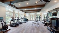 Located at 3510 Corder Dr in Wilmington, NC, this fitness center is a thoughtfully designed single-floor facility that combines functionality with an inviting aesthetic. The space is defined by its striking coffered ceiling accented with dark exposed wood beams, recessed lighting, and multiple ceiling fans that maintain a comfortable atmosphere throughout workouts.

An expansive wall of large windows and glass doors floods the interior with abundant natural light while offering views of lush outdoor greenery and a pool area beyond, creating an energizing environment. The flooring throughout is a durable, neutral-toned carpet tile suited for high-traffic fitness use.

The main workout area is generously proportioned and houses a comprehensive lineup of cardio equipment, including treadmills, ellipticals, a stair climber, and stationary bikes — several branded by TRUE Fitness. A dedicated strength training zone features a multi-functional cable machine, a dumbbell rack with a full range of free weights, and an adjustable workout bench. A large wall-mounted mirror with an integrated display screen enhances the strength area, adding both utility and a modern touch.

A secondary area near the entrance provides a hydration station with a double drinking fountain, a SmartMat exercise guide, and convenient access to restroom facilities, ensuring members have everything needed for a complete and comfortable fitness experience.