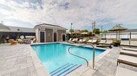 Located at 4664 Kenny Rd in Columbus, Ohio, this outdoor aquatic facility is designed to provide a well-appointed recreational experience. The centerpiece of the space is a generously sized rectangular swimming pool with clear, blue water and mosaic tile detailing along the waterline, adding a polished finishing touch to the pool's interior. Stainless steel entry railings provide safe and convenient access into the water.

The pool deck is finished with light-toned pavers that complement the overall aesthetic and offer a clean, durable surface for foot traffic. A row of cushioned chaise lounges lines the poolside, providing ample seating for relaxation and sunbathing. Additional shaded seating is available further along the deck, furnished with a large umbrella for sun protection.

Adjacent to the pool sits a single-story support building clad in gray lap siding beneath a standing-seam metal roof. The structure provides poolside amenities including restroom facilities, and its traditional architectural detailing integrates cleanly with the surrounding outdoor environment. The entire pool area is enclosed by black metal fencing, ensuring both security and an open, airy feel under open skies.
