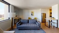 Located at 2625 N Clark Street in Chicago's vibrant Lincoln Park neighborhood, this thoughtfully designed one-bedroom unit offers a seamless blend of modern aesthetics and functional living. The open-concept main living area is anchored by warm hardwood-style flooring and crisp white walls, creating a bright and airy atmosphere. The living room features a tufted gray sofa, a round accent table flanked by chartreuse accent chairs, and a console table with a circular mirror — all accented by contemporary artwork that ties the color palette together. A wall-mounted flat-screen TV and sleek coffee table complete the space. The bedroom is accessed through a striking floor-to-ceiling glass sliding door with a black frame, offering both privacy and visual openness. The bedroom features plush carpeting, a cushioned upholstered headboard, ample natural light from large windows, and city views. The kitchen is well-appointed with two-tone cabinetry — white lower cabinets paired with natural wood upper cabinets — stainless steel appliances including a range, microwave, and dishwasher, and a light tile backsplash. The bathroom offers a clean, modern finish with a wide vanity, large mirror, neutral tile flooring, and a tub-shower combination.