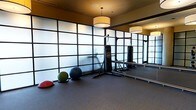 This single-floor fitness studio is thoughtfully designed to support a wide range of training activities. The space is defined by its striking floor-to-ceiling frosted glass partition walls framed in dark metal, which diffuse natural light evenly throughout the room while maintaining a clean, modern aesthetic. Pendant drum light fixtures hang from the warm-toned ceiling, providing soft, ambient illumination that complements the natural light filtering through the glass panels. A large wall-mounted mirror along one side of the room visually expands the space and allows users to monitor their form during workouts. The studio is equipped with a hanging heavy punching bag suspended from a sturdy freestanding rack, making it well-suited for boxing and martial arts training. An assortment of stability and medicine balls in varying sizes are available for functional fitness exercises. The dark rubberized flooring provides excellent traction and cushioning, enhancing both safety and comfort during high-intensity activities. The overall ambiance is focused and professional, striking a balance between an energizing workout environment and a refined, contemporary interior design.