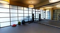 This single-floor fitness studio is thoughtfully designed to support a wide range of training activities. The space is defined by its striking floor-to-ceiling frosted glass partition walls framed in dark metal, which diffuse natural light evenly throughout the room while maintaining a clean, modern aesthetic. Pendant drum light fixtures hang from the warm-toned ceiling, providing soft, ambient illumination that complements the natural light filtering through the glass panels. A large wall-mounted mirror along one side of the room visually expands the space and allows users to monitor their form during workouts. The studio is equipped with a hanging heavy punching bag suspended from a sturdy freestanding rack, making it well-suited for boxing and martial arts training. An assortment of stability and medicine balls in varying sizes are available for functional fitness exercises. The dark rubberized flooring provides excellent traction and cushioning, enhancing both safety and comfort during high-intensity activities. The overall ambiance is focused and professional, striking a balance between an energizing workout environment and a refined, contemporary interior design.