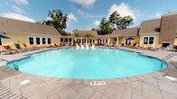 Located at 1500 Parklawn Dr in Charleston, SC, this single-level outdoor amenity space is designed to offer a resort-style experience within a residential community setting. At the heart of the space is a generously sized swimming pool with a smooth, curvilinear shape and a paved stone surround finished in warm earth tones. The pool deck is well-appointed with an abundance of lounge chairs, Adirondack seating, and dining sets shaded by market umbrellas, providing ample space for both relaxing and socializing.

An outdoor fireplace built into a classic brick wall anchors one side of the deck, adding a distinctive architectural focal point and extending the usability of the space into cooler months. A pergola structure adds additional character to the pool surround. Adjacent to the pool area, a covered wraparound porch features white columned railings, a ceiling fan, and a round patio dining table with seating—ideal for enjoying the lush, tree-lined views in a shaded setting. Native South Carolina palmetto palms and mature hardwoods frame the property, providing privacy and a natural backdrop. The clubhouse building itself features cheerful yellow siding with white trim, pitched rooflines, and large windows that reflect the classic Lowcountry architectural aesthetic.