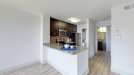 This single-floor space is designed with an open-concept layout that seamlessly connects the living area and kitchen, creating a bright and functional environment. Light hardwood-style flooring runs throughout, complementing the crisp white walls and textured ceilings.

The kitchen is a standout feature, outfitted with rich dark espresso cabinetry that extends to the upper walls, providing generous storage. A stone tile backsplash adds texture and visual interest behind the stainless steel appliances, which include a range, built-in microwave, and a full-sized refrigerator. The peninsula counter is topped with light granite, offering additional prep space and casual seating potential.

The open living area is spacious and unobstructed, with a ceiling fan providing both light and airflow. A pendant light near the entry adds a subtle design detail. The entry door opens directly into this main living space, making the layout feel cohesive and easy to navigate.

A glimpse into an adjacent area suggests an additional room — likely a bathroom or secondary space — with matching dark cabinetry, maintaining a consistent design language throughout the unit.