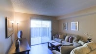 This single-floor space presents a cozy and functional living room as its central feature. The room is bathed in natural light streaming through a large sliding glass door dressed with vertical blinds, which also provides access to an outdoor area. The flooring appears to be a smooth, light-toned surface that complements the warm, neutral wall colors throughout. A textured ceiling adds subtle architectural character to the space. The living room is generously sized to accommodate a plush sectional-style sofa adorned with decorative accent pillows in muted tones of purple and gold, paired with a dark, geometrically designed coffee table and matching ottomans that serve as practical yet stylish centerpieces. Wall art in simple frames punctuates the neutral walls, adding a touch of personality without overwhelming the space. Ambient lighting from a floor lamp in the corner contributes to the warm, inviting atmosphere during evening hours. The overall design aesthetic is casual and comfortable, with a blend of functional furnishings and tasteful décor that creates a relaxed, livable environment well-suited for everyday use.