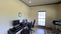 Located at 200-A Heron Cove Dr in Enterprise, Alabama, this single-floor business center is designed to support productivity in a clean, functional environment. The space features a dedicated computer workstation area equipped with multiple desktop computers, keyboards, and a printer, all arranged along a sleek dark desk. Ergonomic leather office chairs provide comfortable seating for extended use. The room is finished with light-toned walls accented by a gray crown molding border that adds a polished, professional touch. Tile flooring throughout contributes to the space's clean and practical aesthetic. A large window fitted with blinds allows natural light to fill the room while maintaining privacy, offering a pleasant view of the surrounding exterior. Recessed ceiling lighting ensures consistent illumination during any hour of the day. A framed piece of wall art adds a subtle decorative element to the otherwise streamlined interior. The overall ambiance is calm, organized, and well-suited for tasks such as printing, browsing, and remote work, making it a highly functional resource for those who need reliable access to computer and office equipment.