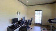 Located at 200-A Heron Cove Dr in Enterprise, Alabama, this single-floor business center is designed to support productivity in a clean, functional environment. The space features a dedicated computer workstation area equipped with multiple desktop computers, keyboards, and a printer, all arranged along a sleek dark desk. Ergonomic leather office chairs provide comfortable seating for extended use. The room is finished with light-toned walls accented by a gray crown molding border that adds a polished, professional touch. Tile flooring throughout contributes to the space's clean and practical aesthetic. A large window fitted with blinds allows natural light to fill the room while maintaining privacy, offering a pleasant view of the surrounding exterior. Recessed ceiling lighting ensures consistent illumination during any hour of the day. A framed piece of wall art adds a subtle decorative element to the otherwise streamlined interior. The overall ambiance is calm, organized, and well-suited for tasks such as printing, browsing, and remote work, making it a highly functional resource for those who need reliable access to computer and office equipment.