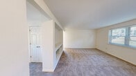 This single-floor residence offers a practical and efficient layout across two bedrooms, a living room, a combined dining room and kitchen, and a connecting hallway. The living room is a standout space, featuring a distinctive built-in shelving unit with glass shelves that serves as a partial room divider, adding both function and architectural character. Neutral-toned carpet runs throughout the living areas and bedrooms, contributing to a clean, cohesive aesthetic.

The kitchen is equipped with warm honey-toned wood cabinetry, dark granite-style countertops, a stainless steel sink, and white appliances including a gas range, dishwasher, and microwave. Tile flooring and open display shelving add practicality to this compact but well-appointed space.

The primary bedroom is generously sized, featuring sliding mirrored closet doors that enhance the sense of space and natural light. The second bedroom is bright and comfortable, with a window overlooking greenery and a standard panel door. The dining area is illuminated by a pendant light fixture and receives natural light through a window with views of the surrounding landscape.

Throughout the residence, white walls and neutral finishes create a bright, airy atmosphere, while windows fitted with horizontal blinds allow for controlled natural light in every room.
