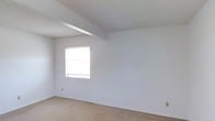 This single-story space presents a straightforward and functional layout with a clean, minimalist aesthetic. The room featured in the image is generously proportioned, offering an open feel enhanced by white-painted walls and a textured ceiling. A structural beam runs across the upper portion of the room, adding a subtle architectural detail to the otherwise understated design. The ceiling varies in height, with one side angling slightly higher, contributing to a sense of openness and volume. A single window fitted with horizontal blinds allows natural light to filter into the space, offering a view of surrounding greenery outdoors. The flooring throughout is covered in neutral-toned carpet, providing a soft and practical surface underfoot. Baseboard vents are visible along the lower walls, indicating a built-in climate control system. The walls are smooth and freshly painted, presenting a blank canvas well-suited for a variety of uses. Overall, the space is clean, vacant, and move-in ready, with a simple and functional character that makes it highly adaptable to different interior arrangements and personal styles.