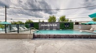 Located at 723 Wheatland St in Phoenixville, Pennsylvania, Immerse is a contemporary outdoor amenity space designed to deliver a resort-like experience. The centerpiece of this thoughtfully designed space is a sparkling pool, framed by a sleek water feature wall bearing the "Immerse" name and flanked by lush vertical greenery that adds a natural, vibrant contrast to the modern hardscape. Glass panel railings border the pool area, providing unobstructed views while maintaining a clean, open aesthetic. The spacious concrete pool deck is generously appointed with neutral-toned chaise lounges and pops of teal through umbrellas and accent furniture, creating a cohesive and inviting color palette. A dedicated lounge zone offers additional seating areas with high-top tables and teal market umbrellas, ideal for social gatherings. Staircase access connects the amenity level to the multi-story building, which features a contemporary gray exterior with large windows overlooking the outdoor space. The overall design balances modern architecture with leisure-focused amenities, making it a well-equipped destination for relaxation and community engagement in the heart of Phoenixville.