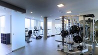 This fitness center offers a well-appointed workout space designed to accommodate a range of exercise needs. The room is defined by its floor-to-ceiling windows along one wall, flooding the space with natural light and providing sweeping panoramic views of the surrounding cityscape — an energizing backdrop for any workout session.

The layout is thoughtfully organized, with dedicated zones for both cardio and strength training. The cardio area features a selection of treadmills, elliptical trainers, and a stationary bike, all positioned to take advantage of the expansive window views. On the opposite side, a strength training station includes a multi-functional weight rack stocked with weight plates and a workout bench, catering to a variety of resistance training exercises.

A central structural column anchors the space visually without impeding functionality. The flooring is a durable, low-profile material suited for high-traffic athletic use. Recessed and panel lighting supplements the natural light, ensuring the space remains bright throughout the day. Large mirrors enhance the sense of openness while allowing users to monitor their form. The overall ambiance is clean, modern, and professionally equipped.