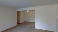 This single-floor space presents a clean, understated interior defined by neutral white walls and wall-to-wall carpeting in a warm beige tone. The layout features an open living area that transitions into a slightly recessed secondary zone, distinguished by a subtle change in ceiling height marked by a structural beam or soffit. This architectural detail creates a natural visual separation between areas while maintaining an open, connected feel throughout the space. The recessed section is equipped with a ceiling-mounted light fixture, providing direct overhead illumination. White painted walls and white trim run consistently throughout, contributing to a bright and cohesive aesthetic. A doorway visible toward the rear left offers access to an additional room or hallway, suggesting further utility beyond the main visible area. The carpeted flooring adds a layer of warmth and softness underfoot. Electrical outlets are positioned along the walls at standard intervals, offering practical functionality. Overall, the space conveys a modest, move-in-ready environment with a straightforward layout suited to comfortable everyday living.