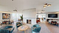 Located in Scottsdale, Arizona, this generously proportioned community living space offers a sophisticated, open-plan layout designed for both relaxed lounging and social gatherings. The interior is anchored by a striking floor-to-ceiling fluted fireplace surround finished in crisp white, serving as an elegant architectural divider between distinct seating areas. Teal leather lounge chairs and a round wood coffee table create an intimate conversation nook, while a variety of dining configurations—featuring warm wood tables and upholstered chairs in earthy tones—provide ample space for communal meals or casual meetings. Natural light pours in through expansive floor-to-ceiling windows and sliding glass doors along multiple walls, offering open views of the surrounding Scottsdale skyline and adjacent outdoor terrace. Light hardwood flooring transitions seamlessly into patterned carpet, delineating the various zones while maintaining visual cohesion throughout. Statement pendant lighting with a mid-century modern aesthetic punctuates the bright white ceiling above. A second lounge area, lined with large glazed walls overlooking a private balcony, features a deep sectional sofa and side tables, ideal for quiet relaxation. The overall ambiance is clean, contemporary, and welcoming—reflecting the vibrant, sun-drenched character of the Arizona desert lifestyle.