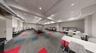 This single-floor facility is designed to support focused, individual work in a clean and organized environment. The open floor plan accommodates a generous number of individual study carrels, each equipped with privacy dividers and chairs, arranged in rows along the walls and across the interior. Additional open seating with white tables and chairs is distributed throughout, offering flexible workspace options for group or casual use.

The interior is finished with neutral gray walls and a drop-panel ceiling fitted with recessed lighting that provides consistent, even illumination throughout the space. Exposed ductwork along one section of the ceiling adds an industrial accent, while structural soffits create subtle visual zones within the open layout.

The flooring is a striking element, featuring a modular carpet tile pattern in a mix of gray, red, and teal, which adds visual energy and helps define areas within the otherwise uniform space. Doors along the perimeter provide access to adjacent utility or support areas. A printer or kiosk station is positioned near one wall, suggesting a functional, service-oriented environment. The overall ambiance is professional, quiet, and purposefully arranged to promote productivity and concentration, making this space well-suited for use as a testing center, library study hall, or academic resource room.