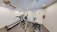 This fitness room is a thoughtfully outfitted exercise space designed to provide residents with a comprehensive workout experience within a compact footprint. The room features clean white walls, dark rubber flooring, and a ceiling fan paired with recessed lighting, ensuring the space remains well-ventilated and properly illuminated during use.

The cardio equipment lineup includes a treadmill with a digital display console, an elliptical cross-trainer, and a recumbent stationary bike, offering a variety of low-impact and high-intensity aerobic options. A full-wall mirror on one side of the room visually expands the space while allowing users to monitor their form during exercise. Positioned in front of the mirror is a three-tier dumbbell rack stocked with a wide range of hex dumbbells, catering to both strength training beginners and more advanced users.

A window with blinds allows natural light to filter into the room, providing a connection to the outdoors. Safety features include a wall-mounted fire extinguisher and a hand sanitizer dispenser near the entry door. The space is reserved for residents only, as noted by posted signage. Overall, this gym delivers a functional and organized environment equipped with the essentials for a well-rounded fitness routine.