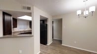 This single-floor space features an open, functional layout that connects the main living area to the kitchen and additional rooms via open doorways and hallways. The living area is finished with warm wood-style laminate flooring and neutral beige walls, creating a clean and understated aesthetic. A textured popcorn ceiling runs throughout, adding a classic residential character, while a ceiling vent ensures comfortable climate control.

The kitchen is positioned adjacent to the main living space and is accessible through an open pass-through counter, offering a semi-open feel. Dark cabinetry lines the kitchen walls, providing a bold contrast against the lighter surroundings. A black full-size refrigerator anchors the kitchen visually, complementing the dark cabinet tones. A sink with a window above it allows for natural light to enter the kitchen area, brightening the workspace.

From the main area, a hallway leads further into the unit, suggesting additional private rooms beyond. White baseboards frame the flooring throughout, adding a crisp finishing detail. The overall ambiance is modest and neutral, offering a practical and move-in-ready environment suited for everyday living.