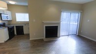 This single-floor space offers a clean, functional layout with notable design elements throughout. The living area serves as the focal point, showcasing a classic white-mantled fireplace set against warm, neutral-toned walls. Rich, dark hardwood flooring spans the space, adding warmth and visual continuity. A large sliding glass door dressed with vertical blinds allows abundant natural light to flood the room, creating a bright and open atmosphere.

The open layout seamlessly connects the living area to the kitchen, which is visible in the background. The kitchen features dark cabinetry paired with light countertops, a sink positioned beneath a window, and dark appliances, offering a functional workspace with its own source of natural light.

The space maintains a neutral color palette throughout — beige walls, white trim, and contrasting dark flooring — lending a cohesive and versatile aesthetic. White baseboards and clean wall finishes add a polished look. Overall, the space balances practicality with comfortable, understated style, making it well-suited for a variety of living arrangements.