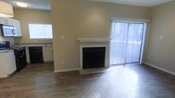 This single-floor space offers a clean, functional layout with notable design elements throughout. The living area serves as the focal point, showcasing a classic white-mantled fireplace set against warm, neutral-toned walls. Rich, dark hardwood flooring spans the space, adding warmth and visual continuity. A large sliding glass door dressed with vertical blinds allows abundant natural light to flood the room, creating a bright and open atmosphere.

The open layout seamlessly connects the living area to the kitchen, which is visible in the background. The kitchen features dark cabinetry paired with light countertops, a sink positioned beneath a window, and dark appliances, offering a functional workspace with its own source of natural light.

The space maintains a neutral color palette throughout — beige walls, white trim, and contrasting dark flooring — lending a cohesive and versatile aesthetic. White baseboards and clean wall finishes add a polished look. Overall, the space balances practicality with comfortable, understated style, making it well-suited for a variety of living arrangements.