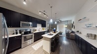 This single-floor residence showcases a thoughtfully designed open-concept layout that seamlessly connects the kitchen, dining, and living areas. The kitchen is a standout feature, equipped with bold dark cabinetry that extends to the ceiling, contrasting sharply against crisp white countertops and a classic tile backsplash. A full suite of stainless steel appliances—including a French door refrigerator, electric range with microwave above, and dishwasher—provides both function and visual appeal. Track lighting and stylish pendant lights suspended over the central island create focused illumination throughout the space. The island offers additional seating and prep space, making it a natural gathering point.

The living area flows naturally from the kitchen, featuring rich wood-style flooring, a ceiling fan, and a large sliding glass door flanked by vertical blinds and drapery panels that admit generous natural light. Decorative wall accents add a contemporary touch to the neutral-toned space.

The bathroom continues the design language of the unit with matching dark cabinetry, dual sinks set into a white countertop, a tall storage tower, and a wide illuminated vanity mirror—offering ample storage and a polished, functional finish.