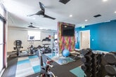 Located at 1200 S Legend Hills Dr in Clearfield, Utah, this fitness center offers a thoughtfully designed exercise room built to support a wide range of workout routines. The space features a colorful, multi-toned carpet tile floor in shades of teal, tan, and charcoal, adding a dynamic visual energy to the room. Large wall-mounted mirrors extend across one side, visually expanding the space and aiding form during exercise. Recessed lighting and ceiling fans with integrated lighting ensure the room remains well-lit and ventilated throughout workouts. Equipment includes a treadmill, elliptical machine, stationary spin bike, and a full dumbbell rack spanning a range of weights. A TRX suspension training system is wall-mounted, offering functional strength training options. Additional accessories such as a BOSU balance trainer, stability ball, and a weight bench round out the versatile setup. A bold accent wall featuring a vibrant geometric mural introduces a modern, energetic aesthetic, complemented by wall-mounted TVs. Natural light filters in through windows and a glass-panel door, keeping the environment bright and inviting. Open shelving provides convenient storage for equipment and personal items, keeping the space organized and accessible.