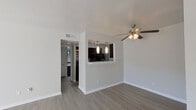 This efficiently laid-out single-story unit offers a thoughtfully designed living space across a compact footprint. The open-concept living and kitchen area forms the heart of the unit, where light gray wood-look flooring runs continuously underfoot, complemented by crisp white walls and clean white baseboards. The living area benefits from a large sliding glass door that floods the space with natural light and provides an outdoor view, while a ceiling fan with integrated lighting adds both function and a subtle design accent.

The kitchen is defined by a raised breakfast bar with a white countertop, functioning as a natural divider between the cooking and living spaces. Dark espresso cabinetry contrasts sharply with the light surroundings, and a mosaic tile backsplash in black and white adds a decorative focal point. Stainless steel appliances, including a microwave, complete the kitchen. Two pendant lights hang above the bar, providing targeted task lighting.

The bedroom is a quiet, well-proportioned room featuring a large window with horizontal blinds that allow for controlled natural light. The bathroom is finished with a dark-framed vanity mirror, black cabinetry with chrome hardware, a white countertop sink, and a tiled shower-tub combination, creating a clean, contemporary aesthetic.