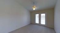 This single-floor, one-bedroom, one-bathroom residence in Arlington (Phase I) offers a clean and functional layout designed for comfortable everyday living. The main living area is finished with neutral wall tones and soft carpeting underfoot, creating a warm and understated atmosphere throughout. A ceiling-mounted light fixture with a decorative medallion detail provides central illumination while adding a subtle architectural accent to the space. One of the most notable features of this room is the set of French doors fitted with privacy blinds, which flood the interior with generous natural light and provide direct access to an outdoor area. The vaulted ceiling angle in the living space contributes to an airy, open feeling, enhancing the sense of volume beyond what the footprint alone might suggest. The overall palette is light and neutral, making the space adaptable to a variety of furnishing styles. The combination of outdoor access, natural light, and simple finishes gives this residence a practical yet pleasant ambiance well suited for single occupancy or a couple.