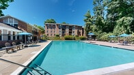 This outdoor amenity space centers around a large, sparkling swimming pool with clearly marked depth indicators, set against a backdrop of mature trees and classic multi-story brick residential buildings. The pool is enclosed by a sleek black metal security fence, providing a sense of privacy and safety for residents. The expansive concrete sundeck surrounds the pool on multiple sides, offering ample room for lounge chairs, Adirondack seating, and umbrella-shaded tables where residents can relax and socialize. Multiple blue market umbrellas are positioned throughout the deck, providing shade during sunny days. A lifeguard stand is stationed poolside, underscoring the community's commitment to safety. The lush green canopy of tall trees frames the perimeter of the space, creating a natural buffer that enhances the serene, resort-like atmosphere. A separate clubhouse structure with a covered porch is visible near the gated entrance to the pool area, suggesting additional community amenities nearby. The well-maintained landscaping, including trimmed hedges and manicured lawns, adds to the polished, welcoming character of this outdoor recreational space.