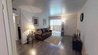 This single-floor unit offers a practical and comfortable layout across several well-defined living spaces. The living room serves as the heart of the home, featuring rich dark hardwood-style flooring, a textured ceiling, and a large sliding glass door that floods the room with natural light and provides direct access to an outdoor patio area. The open feel of the living space transitions seamlessly into a defined dining area, which is neatly tucked into its own alcove and accommodates a round glass-top table with ornate wrought-iron chairs, making it an inviting spot for meals. Framed artwork throughout adds warmth and character to the neutral-toned walls. The kitchen is efficiently designed with dark granite-style countertops, white cabinetry, and black appliances, including a full-size refrigerator. The bedroom is a calm and restful retreat, finished with plush carpeting, a ceiling fan with integrated lighting, and a window that allows in soft natural light. The bathroom continues the dark countertop aesthetic with a granite-style vanity surface, dual mirrors above the sink, and clean white fixtures. Overall, the space presents a cohesive and functional design with a neutral palette throughout.