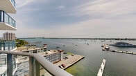 This single-floor waterfront space is defined by its outstanding outdoor vantage point. The private balcony, enclosed by a sleek metal railing, extends the living experience outward and offers unobstructed, panoramic views across a broad bay. From this elevated position, the scenery encompasses an active marina with sizable yachts docked at nearby piers, as well as dozens of sailboats and motorboats dotting the open water. The distant shoreline and city skyline stretch across the horizon beneath a wide, open sky, lending the space a sense of expansiveness that is rare and highly desirable. The waterfront setting provides a dynamic backdrop that shifts with the light and activity on the bay throughout the day. While the interior images do not provide sufficient detail to describe the indoor spaces, the overall character of this unit is strongly shaped by its elevated position and its direct visual connection to the surrounding waterway. The balcony serves as a key architectural feature, seamlessly blending indoor and outdoor living and making the bay views the central, defining element of the entire space.
