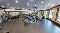 This fitness center is a spacious, single-floor facility designed to support a comprehensive workout experience. The room is anchored by a long row of cardio machines, including ellipticals and treadmills from Freedom and similar brands, arranged in orderly lines across a durable rubber flooring that provides both safety and comfort underfoot. A dedicated free weights area lines one wall, featuring a full rack of dumbbells alongside weight benches, catering to strength training needs. Large mirrors span a significant portion of the walls, creating a sense of openness while allowing users to monitor their form. Recessed lighting is distributed evenly across the ceiling, ensuring the space is consistently well-lit throughout. A ceiling fan adds to the ventilation and comfort of the room. Natural light filters in through windows along one side of the facility, contributing to a bright and motivating atmosphere. The neutral color palette of the walls and flooring gives the space a clean, professional aesthetic. Overall, the fitness center is thoughtfully arranged to maximize usable floor space while accommodating a variety of exercise equipment suitable for both cardio and strength training routines.