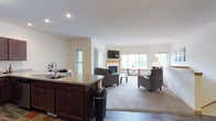This single-floor residence is designed around an open-concept layout that effortlessly connects the kitchen, dining, and living areas into one cohesive, flowing space. The kitchen features a dark-stained wood island with a built-in sink and ample cabinetry, providing generous counter space and storage. Hardwood flooring anchors the kitchen area, while plush carpeting defines the expansive living room beyond. The living room is the centerpiece of the home, highlighted by a corner fireplace with a light-toned surround, serving as a warm and inviting focal point. A wall-mounted television above the fireplace adds a modern touch, while comfortable seating arrangements create a relaxed, functional atmosphere. Large windows along multiple walls flood the space with natural light, offering views of the surrounding greenery and lending an airy, open feel to the interior. The neutral color palette throughout — soft whites on the walls and ceilings paired with warm wood tones — creates a clean, welcoming ambiance. Recessed lighting complements the natural illumination, ensuring the space is well-lit at any hour. Overall, this home offers a practical yet comfortable living environment with a thoughtful, open design.
