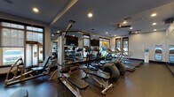 This fitness center is a comprehensively outfitted single-level facility designed to support a full range of workout routines. The space is finished with durable rubber flooring throughout, providing a safe and functional surface suited to both cardio and strength training activities. Recessed lighting, paired with ceiling fans, ensures the room remains well-lit and well-ventilated during use.

The cardio section features multiple Star Trac treadmills and elliptical machines, each equipped with digital displays. These are positioned near large windows fitted with roller shades, allowing natural light to filter in while maintaining privacy. The views to the surrounding outdoor area lend an open, airy quality to this zone.

The strength training area includes a multi-station cable machine, adjustable benches, and a selection of selectorized weight equipment. Floor-to-ceiling mirrors line one wall, visually expanding the space and allowing users to monitor their form. Wall-mounted flat-screen televisions are positioned throughout for entertainment during workouts.

Additional amenities include a water cooler station, a towel and accessory rack stocked with resistance equipment and exercise balls, and a paper towel dispenser. A set of glass double doors serves as the main entry point, with posted fitness center rules displayed prominently nearby. The overall design is clean, functional, and well-maintained.