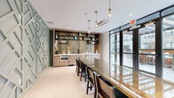 Located at 308 NJ-31 N in Flemington, New Jersey, this elegantly designed meeting and dining room offers a refined and functional space for gatherings and collaborative use. The room is defined by a striking gold hexagonal mosaic tile backsplash that anchors the built-in bar area, complete with light wood cabinetry, open shelving, and an under-counter appliance — an ideal setup for catered events or casual entertaining. A sculptural sage-green geometric accent wall adds a sophisticated architectural focal point, featuring an intricate lattice pattern that introduces texture and visual depth. At the center of the room sits a long glass-top conference table surrounded by cushioned chairs, providing ample seating for meetings or dining. Pendant lights with brass fixtures hang above, casting warm, ambient light across the space. Large floor-to-ceiling windows and glass doors line the exterior wall, flooding the interior with natural light and offering views of an adjacent outdoor courtyard and the surrounding building complex. Large-format light tile flooring and glass partition walls further enhance the open, airy feel of the space. Potted plants positioned near the windows add a touch of greenery, contributing to a welcoming and professional atmosphere throughout.