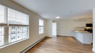 Located at Carrier Woods in Scarborough, Maine, this single-floor residence offers a well-designed and inviting living environment. The main living area is highlighted by warm hardwood flooring that extends throughout the space, contributing to a bright and welcoming atmosphere. Large windows with white blinds allow generous amounts of natural light to fill the room, enhancing the open and airy feel of the interior. The walls are finished in a soft, neutral cream tone that complements the light wood flooring and creates a clean, cohesive aesthetic. A baseboard heating system runs along the exterior wall, ensuring comfortable warmth throughout the colder Maine seasons. The space flows naturally from the main living area toward an entry door and an adjacent kitchen area, visible in the background, where light countertops add to the home's fresh, modern appeal. Flush-mount ceiling lighting and a smoke detector are cleanly integrated into the white ceiling. The overall design is straightforward and functional, with a neutral palette and quality finishes that make the space adaptable to a variety of interior styles. The exterior views through the windows reveal neighboring structures, situating this residence within a residential community setting.