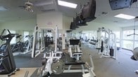 This spacious fitness center is designed to accommodate a comprehensive workout routine, offering an impressive array of equipment across a single open floor. The layout is thoughtfully organized, with strength training machines and free weight stations positioned throughout the room for ease of movement and access. A dedicated dumbbell rack lines one side of the facility, offering a full range of weights for varied strength training exercises. Multiple weight machines with adjustable settings cater to both beginners and experienced gym-goers alike. Overhead, ceiling-mounted flat-screen televisions are strategically placed throughout the space, allowing members to stay entertained during cardio and strength sessions. The facility benefits from large windows that invite natural light, creating a bright and energizing atmosphere. Recessed and panel lighting supplements the natural light for consistent illumination throughout. Light-colored walls and mirrored surfaces visually expand the space, adding to the open, airy feel of the gym. The flooring transitions between rubber and carpet zones, providing appropriate surfaces for both heavy lifting and machine-based workouts. Vaulted ceiling sections add a sense of height and spaciousness to the overall environment.