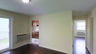 This single-floor space offers a straightforward, functional layout designed for comfortable everyday living. The main living area is finished with dark hardwood flooring that contrasts cleanly against light yellow-toned walls and white baseboards, creating a bright and neutral interior palette. A flush-mount ceiling light provides overhead illumination, while a wall-mounted PTAC unit along the lower wall offers independent climate control.

An open passthrough connects the living area directly to the kitchen, which features rich medium-toned wood cabinetry on both the upper and lower sections, providing ample storage space. The kitchen's tiled flooring distinguishes it from the hardwood in the main living area, and light-colored countertops complement the warm wood tones throughout.

A doorway visible on the right side of the main room suggests access to additional areas of the unit, hinting at further functional spaces beyond the main living and kitchen zones. The overall ambiance is clean and understated, with a simple, practical design that prioritizes utility and ease of maintenance.