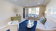This single-floor space offers a well-designed interior that combines comfort and style in a cohesive, contemporary aesthetic. The living area serves as the focal point of the space, featuring recessed ceiling lighting and smooth, light-toned walls that create a clean and airy atmosphere. A large multi-pane window dominates one wall, flooding the room with natural light and offering views of the surrounding greenery and neighboring buildings.

The living area is anchored by plush white seating arranged around a sleek oval glass coffee table with a chrome frame, set atop a rich navy blue area rug that adds a bold pop of color to the neutral palette. A decorative sunburst mirror with a starburst frame adds an artistic touch to one wall, while a sage green carved wooden credenza introduces both storage and a warm, eclectic accent. Decorative vases and greenery further enhance the inviting ambiance.

Adjacent to the main seating area, a compact glass-top dining set with upholstered chairs is positioned near the window, making the most of the natural light for a functional yet elegant dining nook. Light hardwood-style flooring runs throughout, complementing the overall bright and cohesive design. The space balances practicality with thoughtful décor, resulting in a comfortable and visually appealing environment.