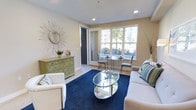 This single-floor space offers a well-designed interior that combines comfort and style in a cohesive, contemporary aesthetic. The living area serves as the focal point of the space, featuring recessed ceiling lighting and smooth, light-toned walls that create a clean and airy atmosphere. A large multi-pane window dominates one wall, flooding the room with natural light and offering views of the surrounding greenery and neighboring buildings.

The living area is anchored by plush white seating arranged around a sleek oval glass coffee table with a chrome frame, set atop a rich navy blue area rug that adds a bold pop of color to the neutral palette. A decorative sunburst mirror with a starburst frame adds an artistic touch to one wall, while a sage green carved wooden credenza introduces both storage and a warm, eclectic accent. Decorative vases and greenery further enhance the inviting ambiance.

Adjacent to the main seating area, a compact glass-top dining set with upholstered chairs is positioned near the window, making the most of the natural light for a functional yet elegant dining nook. Light hardwood-style flooring runs throughout, complementing the overall bright and cohesive design. The space balances practicality with thoughtful décor, resulting in a comfortable and visually appealing environment.
