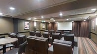 This private screening room is designed to deliver a true cinematic experience in a comfortable and well-appointed setting. The space is arranged with rows of plush, dark leather recliner chairs positioned in a tiered layout, ensuring clear sightlines to the large projection screen mounted at the front of the room. A ceiling-mounted projector provides high-quality image projection, making the room fully equipped for movie screenings, presentations, or private events.

The interior design draws clear inspiration from classic movie theaters. Warm, neutral-toned walls are complemented by rich brown draping that frames the screen and adds a sense of depth and elegance to the space. Recessed ceiling lighting, along with subtle wall sconces, creates a warm and inviting ambiance that can be adjusted to suit the mood of any screening.

Classic black-and-white movie posters in framed displays adorn the walls, reinforcing the cinematic theme and adding a touch of vintage Hollywood charm. The drop ceiling with acoustic tiles contributes to sound management, enhancing the overall audio experience within the room. The combination of thoughtful design, quality seating, and professional-grade equipment makes this screening room a standout venue for entertainment and private gatherings.