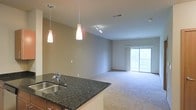 This single-floor space is designed with an open-concept layout that seamlessly connects the kitchen and living areas. The kitchen features dark granite countertops with a stainless steel sink, complemented by elegant pendant lighting that hangs above the counter, creating a warm and functional workspace. The kitchen's open design allows for a clear sightline into the adjacent living area, making it ideal for entertaining or everyday living.

The living area is finished with neutral-toned carpeting that adds warmth and comfort underfoot. A track lighting fixture installed on the ceiling provides flexible illumination throughout the space. The neutral-toned walls throughout create a clean, versatile backdrop that suits a variety of interior styles.

A standout feature of the living area is the sliding glass door, which floods the space with natural light and provides direct access to an outdoor balcony. The view through the glass doors hints at a green, outdoor setting, enhancing the overall sense of openness. The combination of natural light, modern lighting fixtures, and the flowing layout gives the space a bright and welcoming ambiance.