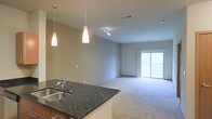 This single-floor space is designed with an open-concept layout that seamlessly connects the kitchen and living areas. The kitchen features dark granite countertops with a stainless steel sink, complemented by elegant pendant lighting that hangs above the counter, creating a warm and functional workspace. The kitchen's open design allows for a clear sightline into the adjacent living area, making it ideal for entertaining or everyday living.

The living area is finished with neutral-toned carpeting that adds warmth and comfort underfoot. A track lighting fixture installed on the ceiling provides flexible illumination throughout the space. The neutral-toned walls throughout create a clean, versatile backdrop that suits a variety of interior styles.

A standout feature of the living area is the sliding glass door, which floods the space with natural light and provides direct access to an outdoor balcony. The view through the glass doors hints at a green, outdoor setting, enhancing the overall sense of openness. The combination of natural light, modern lighting fixtures, and the flowing layout gives the space a bright and welcoming ambiance.