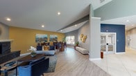 Located at 14901 E Mississippi Ave in Aurora, Colorado, this single-floor communal space offers a well-appointed, open-concept layout designed for both relaxation and social interaction. The interior is defined by vaulted ceilings that lend a sense of spaciousness, complemented by recessed lighting and light wood-plank flooring throughout.

The lounge area serves as the focal point of the space, anchored by a striking corner fireplace clad in dark stacked stone with a white mantel and a decorative sunburst mirror above. Comfortable seating, including a light gray sofa and navy accent chairs, is arranged around a wood coffee table atop a patterned area rug. Large windows allow natural light to fill the room while offering views of the surrounding outdoor greenery.

Adjacent to the lounge, a dedicated dining area features walnut-toned tables and navy upholstered chairs, positioned near additional windows. A seating nook along one wall is accented by a sculptural gold metallic wall installation, adding an artistic element to the space.

A kitchenette area provides practical amenities, including a stainless steel sink, countertop space, floating wood shelves, and a coffee station, all set against a pearlescent white tile backsplash. The overall color palette of warm gold, cool gray, and navy blue creates a cohesive and inviting atmosphere throughout.