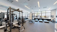 Frankford Fitness is a thoughtfully designed exercise facility located at 1120 Frankford Avenue in Philadelphia, PA. The single-floor space features a contemporary aesthetic with clean white walls, recessed linear lighting, and durable carpet flooring throughout, creating a bright and motivating environment for exercise.

The facility is comprehensively equipped for both cardio and strength training. A row of Star Trac treadmills lines the floor-to-ceiling glass curtain wall, which floods the space with natural light and offers views of the streetscape and outdoor courtyard beyond. This glazed façade gives the gym an open, airy feel that distinguishes it from typical enclosed fitness spaces.

The strength training area includes a Body-Solid multi-station cable machine, a full dumbbell rack with a wide range of weights, adjustable benches, and a squat/power rack. Additional equipment includes a spin bike, slam balls, resistance bands, and various functional fitness accessories. A wall-mounted flat-screen TV is positioned above a decorative geometric-patterned accent wall, providing an engaging focal point for the training floor.

Full-length mirrors along one wall visually expand the space and assist with form during workouts. The entrance, marked by a branded "Frankford Fitness" sign on a textured stone accent wall, leads directly to the main training floor, with a storage cubby unit providing space for personal belongings. The facility presents a polished, modern fitness experience.