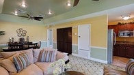This single-floor space exudes a warm and welcoming character, brought to life through a thoughtfully chosen color palette and classic design elements. The walls are dressed in a soft, sunny yellow on the upper half, transitioning to a muted sage green on the lower half, divided by crisp white wainscoting that adds a polished, traditional touch throughout. The ceiling mirrors the sage green tone, creating a cohesive and enveloping ambiance, while recessed lighting and ceiling fans ensure both comfort and functionality. The living area is anchored by a plush sofa adorned with striped accent pillows, complemented by a decorative floral arrangement that adds a fresh, natural element to the space. Underfoot, a patterned area rug in muted tones ties the room together with subtle elegance. Dark-stained louvered closet doors provide a striking contrast against the lighter wall tones, adding depth and visual interest. A solid white door leads to an adjoining room, maintaining the clean, bright aesthetic. The overall ambiance is relaxed yet refined, making this a comfortable and stylish retreat that balances casual living with classic design sensibilities.